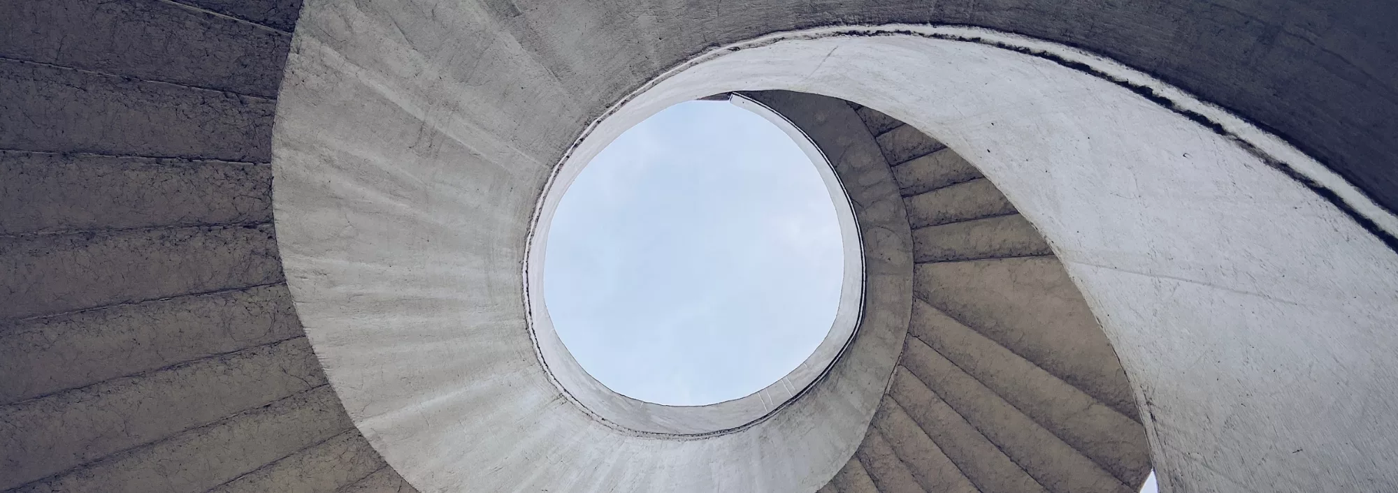 Looking up at the sky through a modern concrete stairwell, an almost abstract pattern of concentric or winding circles, for Marksman Wealth Management's theme of being focused on the clients' goals.